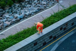 City gardener in orange uniform tending to plants on a stone wall in a park.
